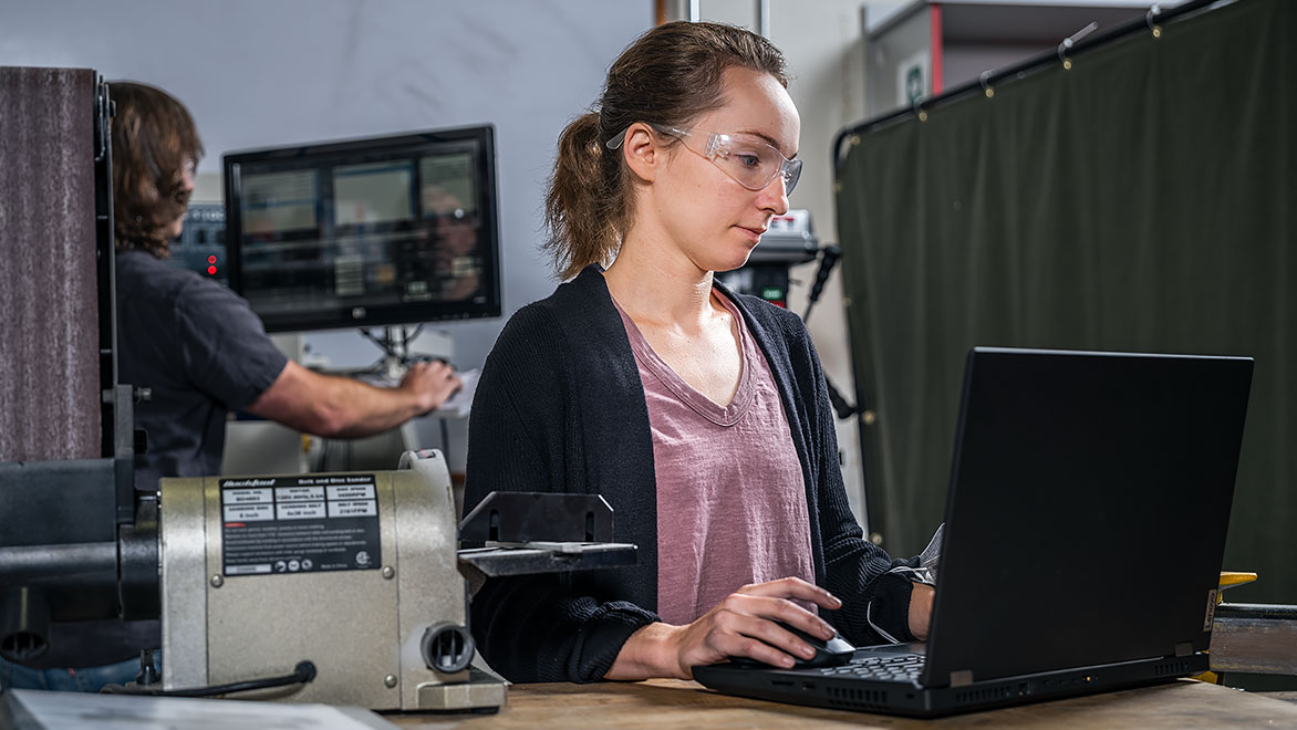 Woman wearing safety glasses works on laptop in workplace